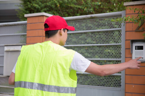 Personne en gilet jaune devant un portail extérieur d'une maison, en train de sonner pour entrer